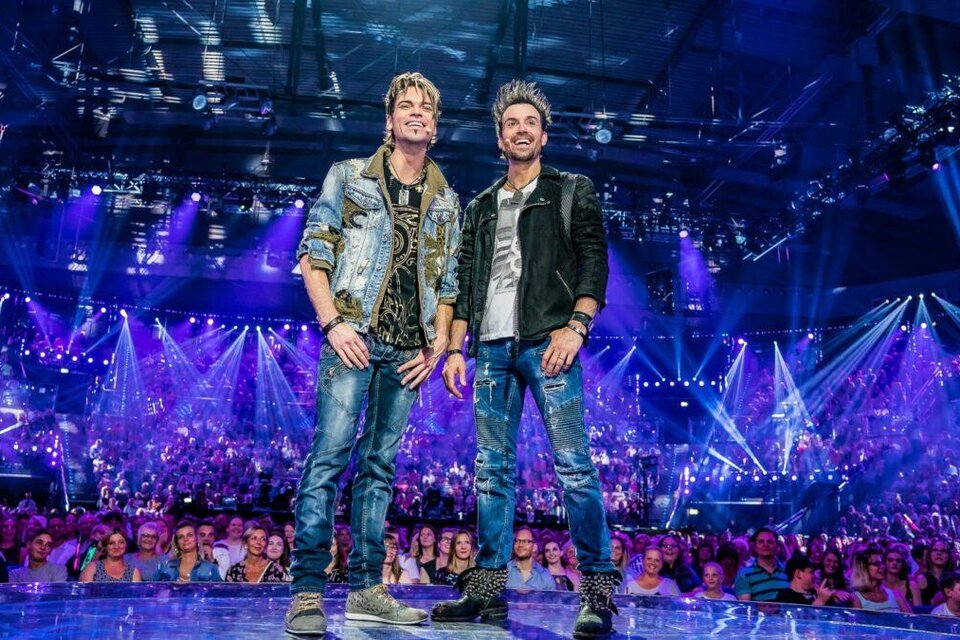 Zwei Musiker stehen auf einer Bühne mit bunten Lichtern und einem begeisterten Publikum. Die Atmosphäre ist festlich und energetisch. | © ralph-larmann-gross