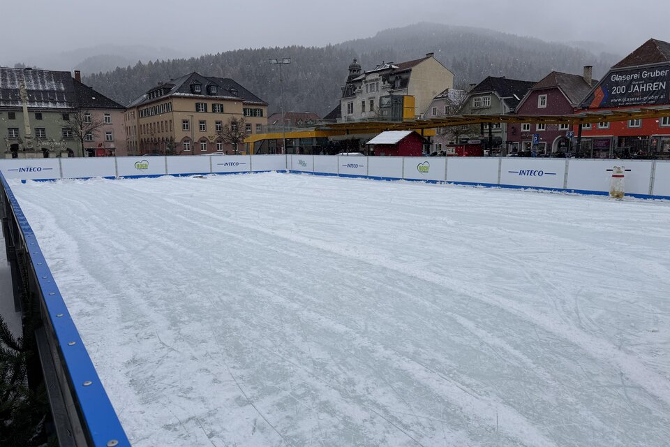 Eine leere Eisbahn mit einer glatten Oberfläche und einem winterlichen Hintergrund. Die Umgebung zeigt Häuser und eine neblige Bergkulisse. | © TV Hochsteiermark