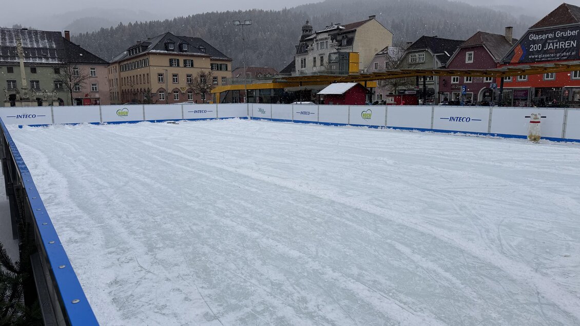 Eine leere Eisbahn mit einer glatten Oberfläche und einem winterlichen Hintergrund. Die Umgebung zeigt Häuser und eine neblige Bergkulisse. | © TV Hochsteiermark