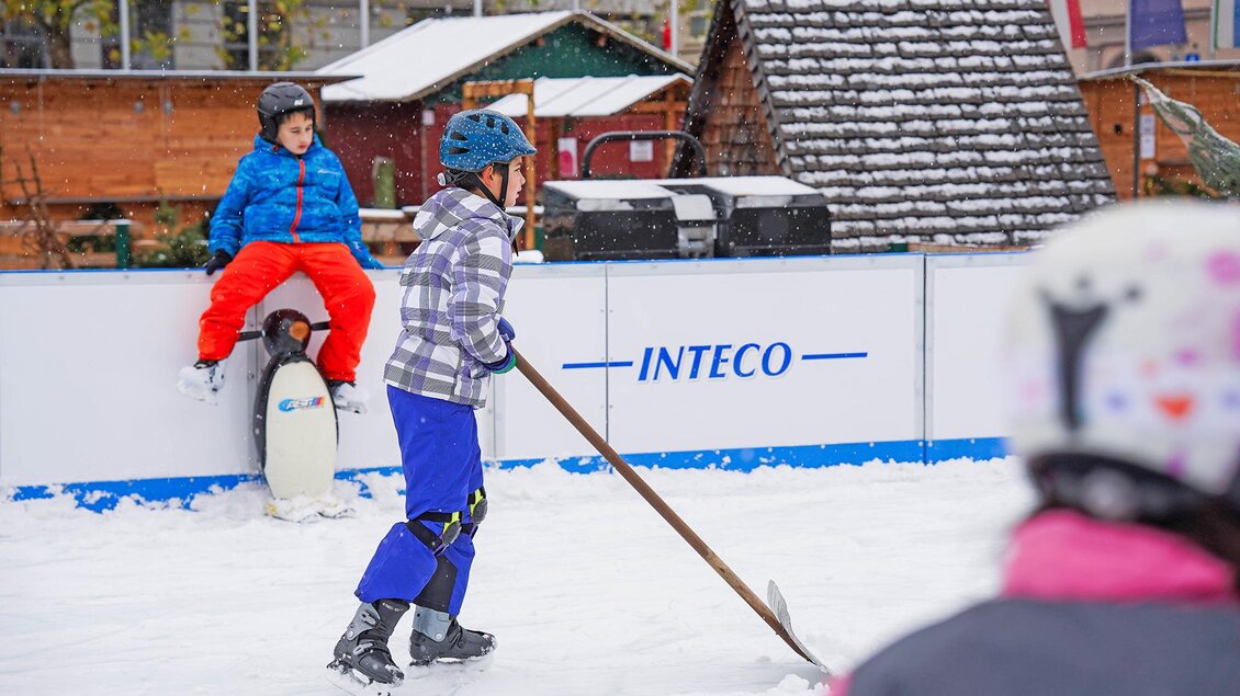 Kinder spielen auf einer Kunsteisbahn. Ein Junge steht mit einem Besen und ein anderer sitzt auf der Bande. | © StadtBruck, Vassilev