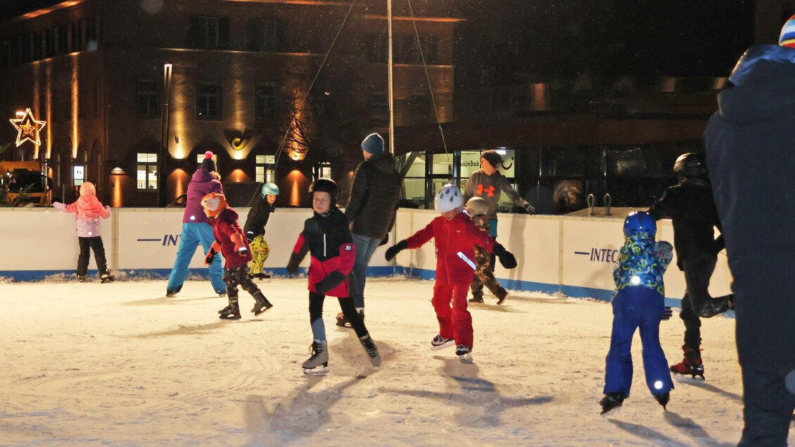 Eine Gruppe von Kindern fährt fröhlich Schlittschuh auf einer beleuchteten Eisbahn. Im Hintergrund sind dekorierte Gebäude und Weihnachtslichter zu sehen. | © StadtBruck, Vassilev