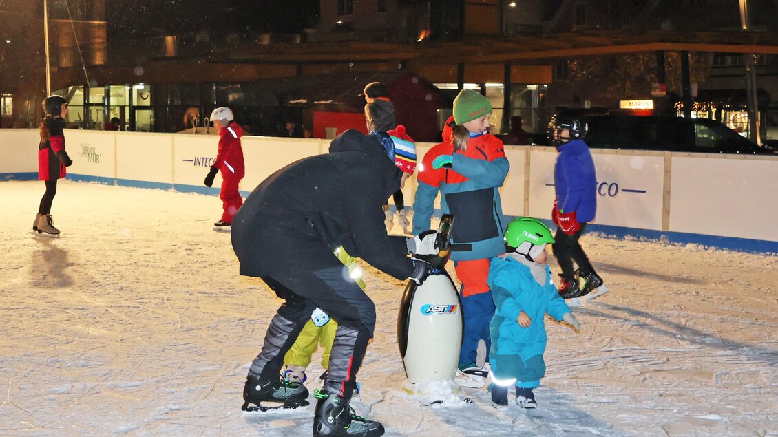 Eine Eislauffläche, auf der Kinder mit Hilfe von Erwachsenen Schlittschuh laufen. Es ist Nacht, und die Szene ist festlich beleuchtet. | © StadtBruck, Vassilev