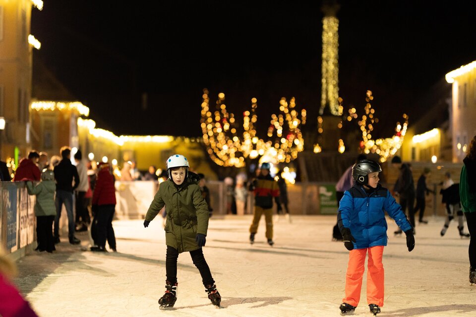 Eine Eislaufbahn bei Nacht mit vielen fröhlichen Menschen. Kinder skaten in bunten Jacken unter funkelnden Lichtern.