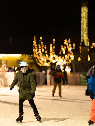 An ice skating rink at night with many cheerful people.  
Children skate in colorful jackets under sparkling lights.