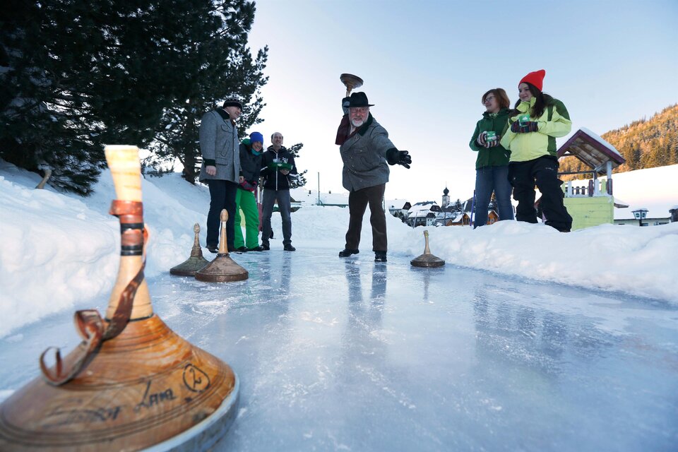 Eine Gruppe von Menschen spielt im Schnee eine traditionelle Eissportart. Im Hintergrund sind Bäume und eine bewirtete Hütte zu sehen. | © Herbert Raffalt