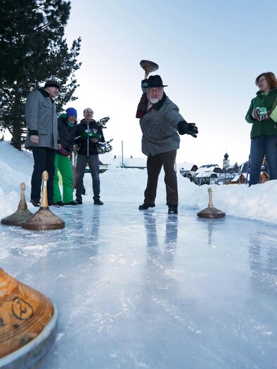 A group of people is playing a traditional ice sport in the snow. In the background, trees and a hosted cabin can be seen. | © Herbert Raffalt