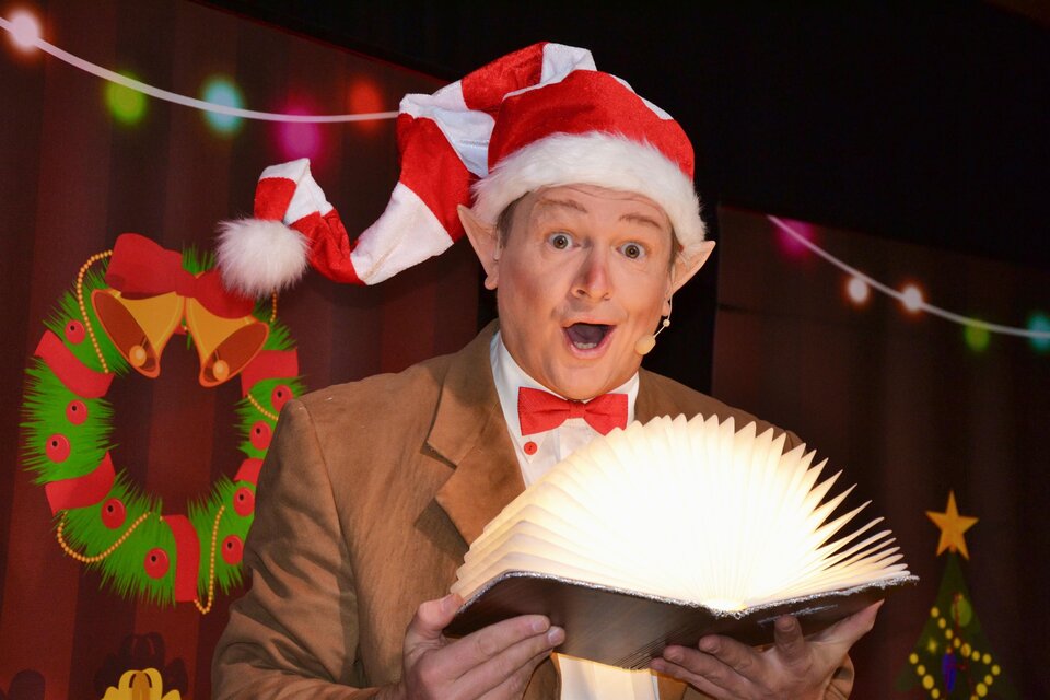 Ein fröhlicher Mann in einem Weihnachtsmann-Hut liest aus einem großen, beleuchteten Buch. Im Hintergrund sind festliche Dekorationen mit Weihnachtsmotiven zu sehen. | © Stadtgemeinde Trofaiach