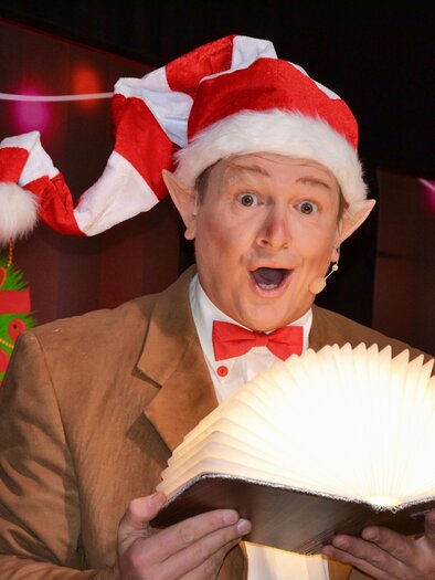 A cheerful man in a Santa hat reads from a large, illuminated book. In the background, festive decorations with Christmas motifs are visible. | © Stadtgemeinde Trofaiach