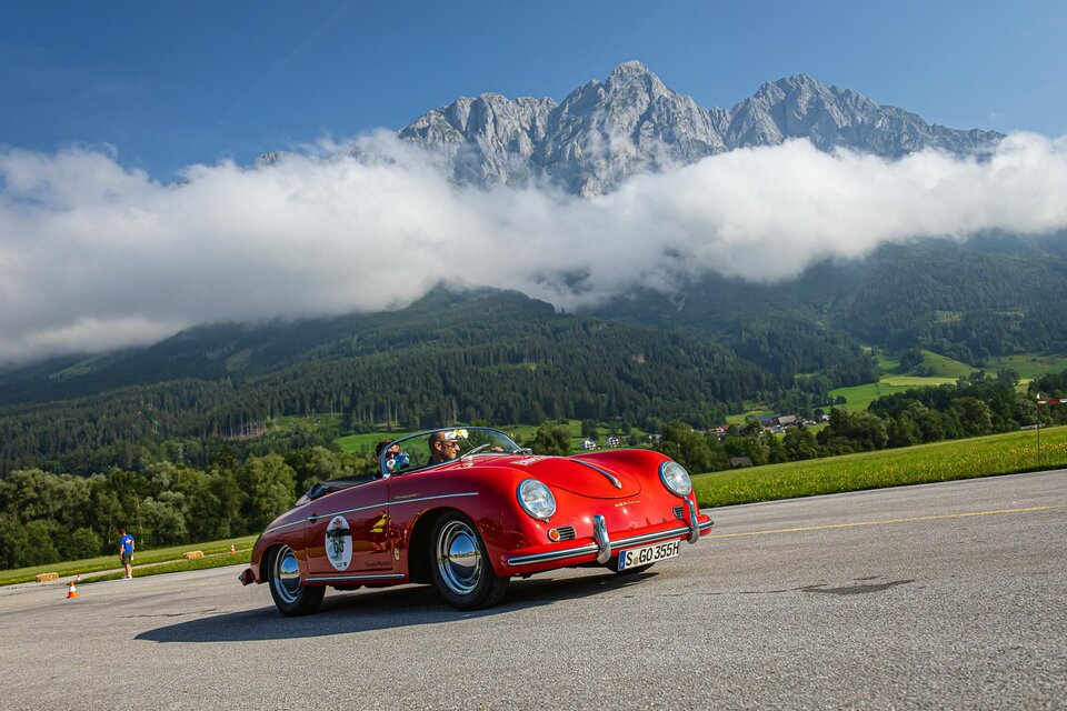 Ein rotes Sportauto fährt vor einer beeindruckenden Berglandschaft. Die Wolken umgeben die Gipfel, während die grüne Wiese im Vordergrund zu sehen ist. | © Steering Media