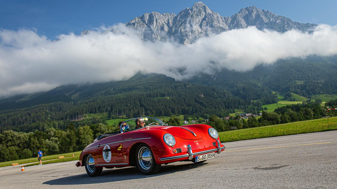 Ein rotes Sportauto fährt vor einer beeindruckenden Berglandschaft. Die Wolken umgeben die Gipfel, während die grüne Wiese im Vordergrund zu sehen ist. | © Steering Media