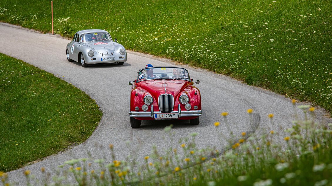 Zwei Oldtimer fahren auf einer kurvenreichen Landstraße. Die grüne Landschaft im Hintergrund verleiht der Szene einen malerischen Charme. | © Peter Meierhofer
