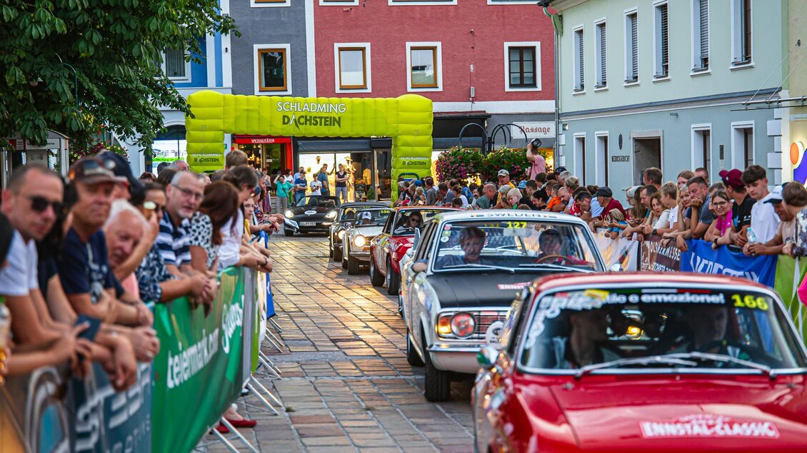 Eine lebhafte Straßenveranstaltung mit klassischen Autos, die an einer großen Menschenmenge vorbeifahren. Im Hintergrund sind bunte Gebäude und ein gelbes Zelt zu sehen. | © Steering Media