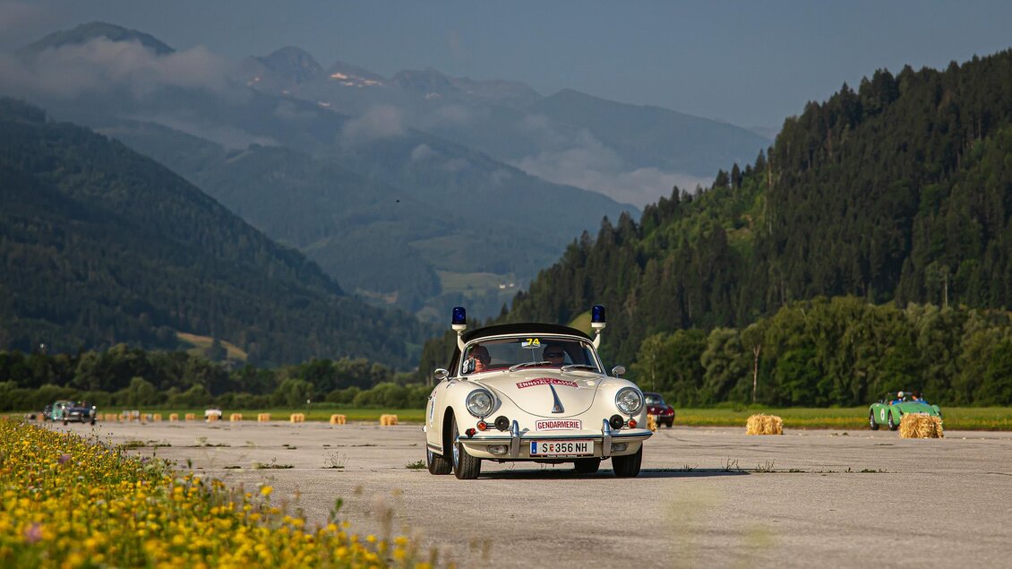 Ein klassisches Auto fährt auf einer Straße umgeben von Blumen und Bergen. Der Himmel ist klar und die Landschaft wirkt idyllisch. | © Steering Media
