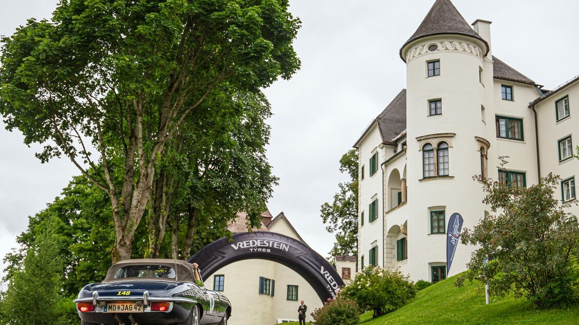 Ein traditionelles Schloss mit einem großen, grünen Rasen und hohen Bäumen. Im Vordergrund sieht man ein klassisches Auto, das auf den Eingang zum Schloss zufährt. | © Steering Media