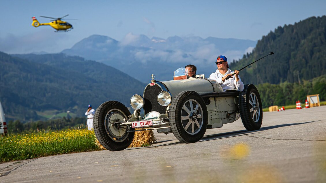 Ein klassisches Auto fährt auf einer Straße mit Alpenlandschaft im Hintergrund. Ein Hubschrauber fliegt darüber, während die Sonne scheint. | © Steering Media