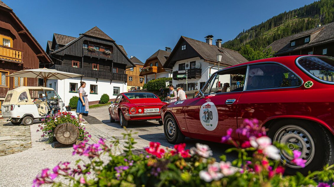 Ein historisches Autorennen in einer malerischen Stadt mit alten Holzhäusern. Blumen und Menschen sind im Vordergrund sichtbar. | © Steering Media