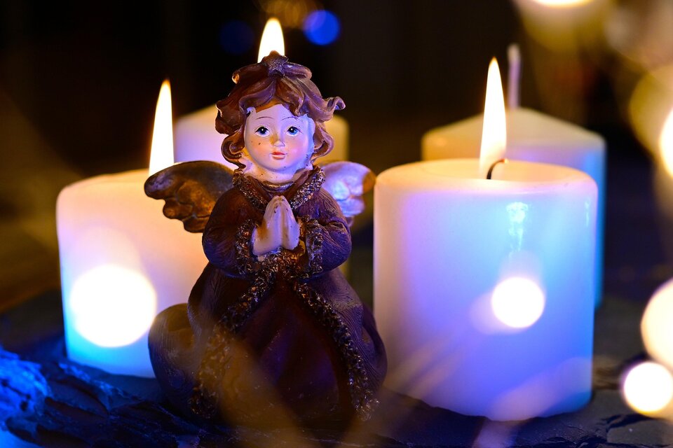 Eine kleine Engelfigur sitzt zwischen drei brennenden Kerzen. Sanftes Licht erhellt die Szene und schafft eine ruhige Atmosphäre. | © pixabay.com