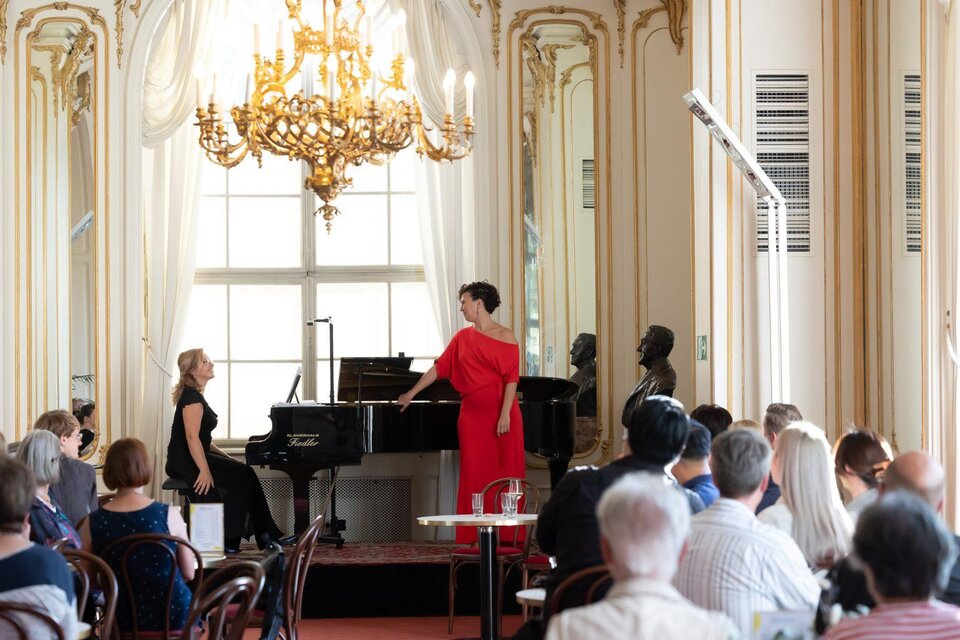 Eine elegante Aufführung in einem stilvollen Raum mit Kronleuchtern und großen Fenstern. Zwei Sängerinnen stehen am Klavier und unterhalten ein Publikum. | © Oper Graz