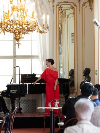 Eine elegante Aufführung in einem stilvollen Raum mit Kronleuchtern und großen Fenstern. Zwei Sängerinnen stehen am Klavier und unterhalten ein Publikum. | © Oper Graz