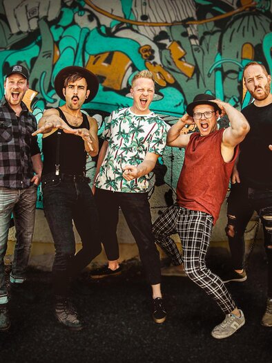 Eine Gruppe von fünf Männern posiert vor einer bunten Graffiti-Wand. Sie zeigen fröhliche und energiegeladene Gesten. | © (c)Faeaschtbaenkler