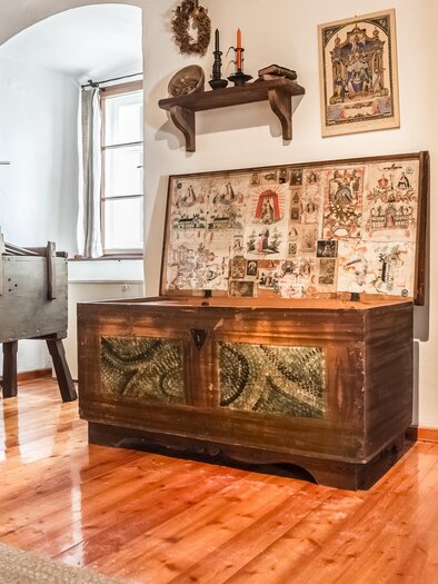 Ein gemütlicher Raum mit rustikaler Einrichtung. Im Vordergrund steht eine verzierte Holztruhe und im Hintergrund sind weitere Möbel und Dekorationen zu sehen. | © TV Hochsteiermark, Fred Lindmoser