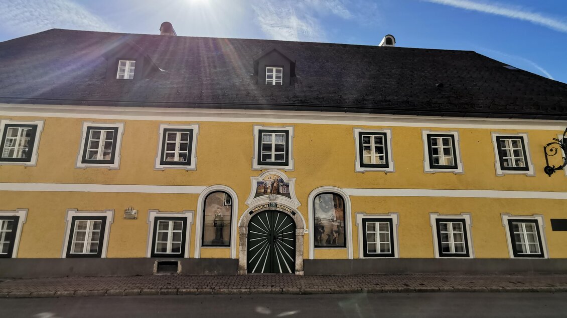 Ein gelbes Gebäude mit mehreren Fenstern und einer großen, grünen Tür. Der Himmel ist klar und die Sonne scheint. | © TV Hochsteiermark