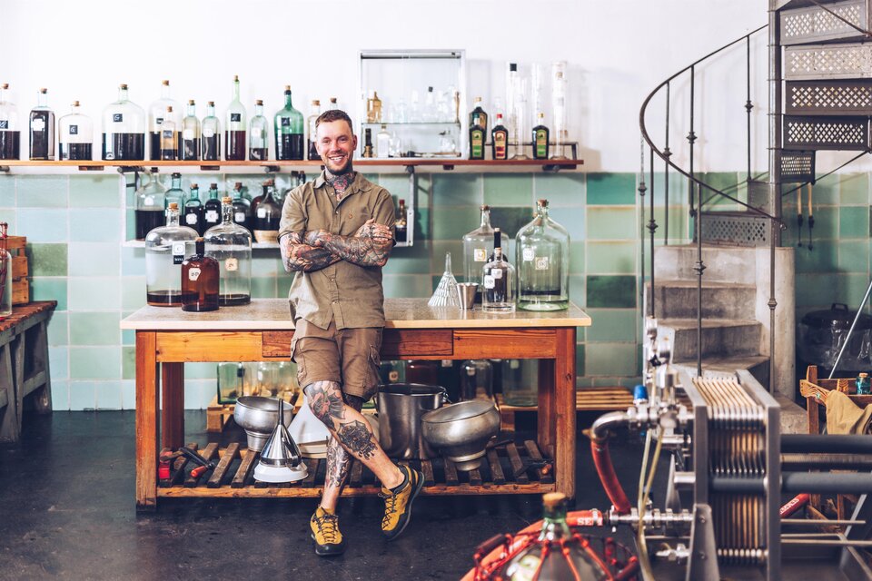Ein lächelnder Mann steht an einer großen Holztheke in einer modernen Brauerei. Im Hintergrund sind viele Flaschen und Glasschalen sichtbar. | © Arzberger Likörmanufaktur
