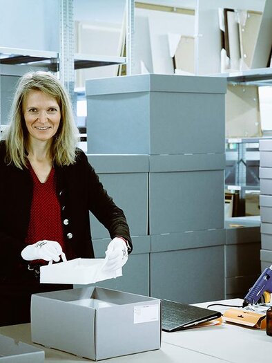 A smiling woman stands at a table in a storage room and holds a white box. In the background, many gray cartons and shelves with additional boxes are visible. | ©  Foto Freisinger
