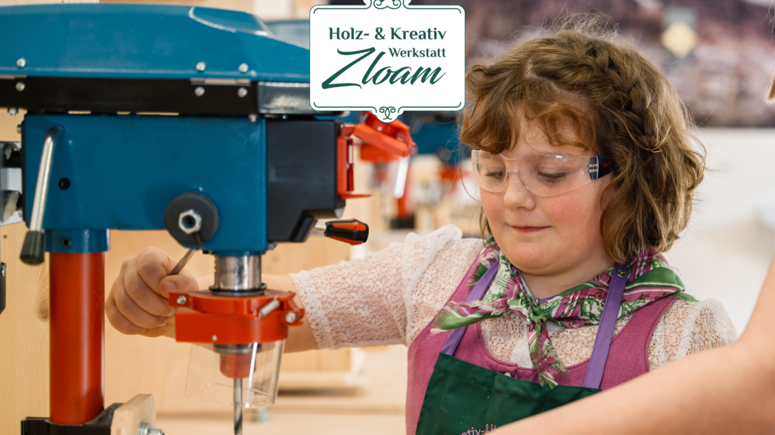 Ein Mädchen arbeitet an einer Maschine in einer Holzwerkstatt. Sie trägt eine Schutzbrille und lächelt, während sie konzentriert ist. | © Narzissendorf Zloam, www.zloam.at