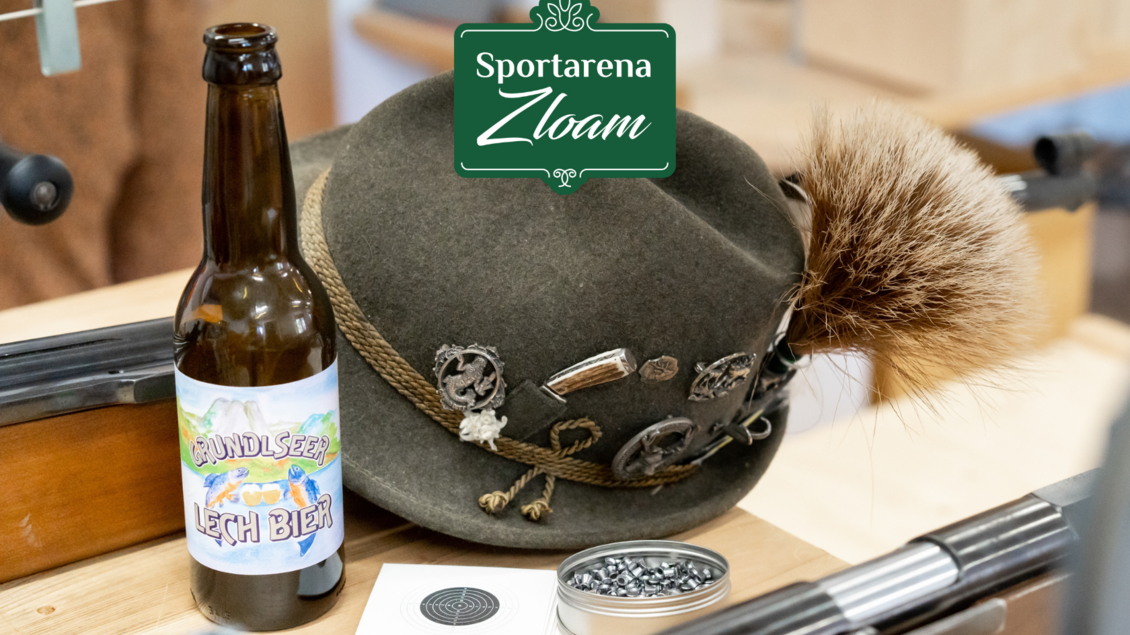 Ein traditioneller bayerischer Hut mit einer Feder und ein Bier mit dem Etikett "Lech Bier" stehen auf einem Tisch. Im Hintergrund ist das Logo von "Sportarena Zloam" zu sehen. | © Narzissendorf Zloam, www.zloam.at