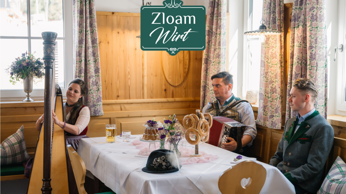 Ein gemütliches Speiselokal mit traditioneller Dekoration. Musizierende Gäste, die Harfe und Akkordeon spielen, während ein Tisch festlich gedeckt ist. | © Narzissendorf Zloam, www.zloam.at