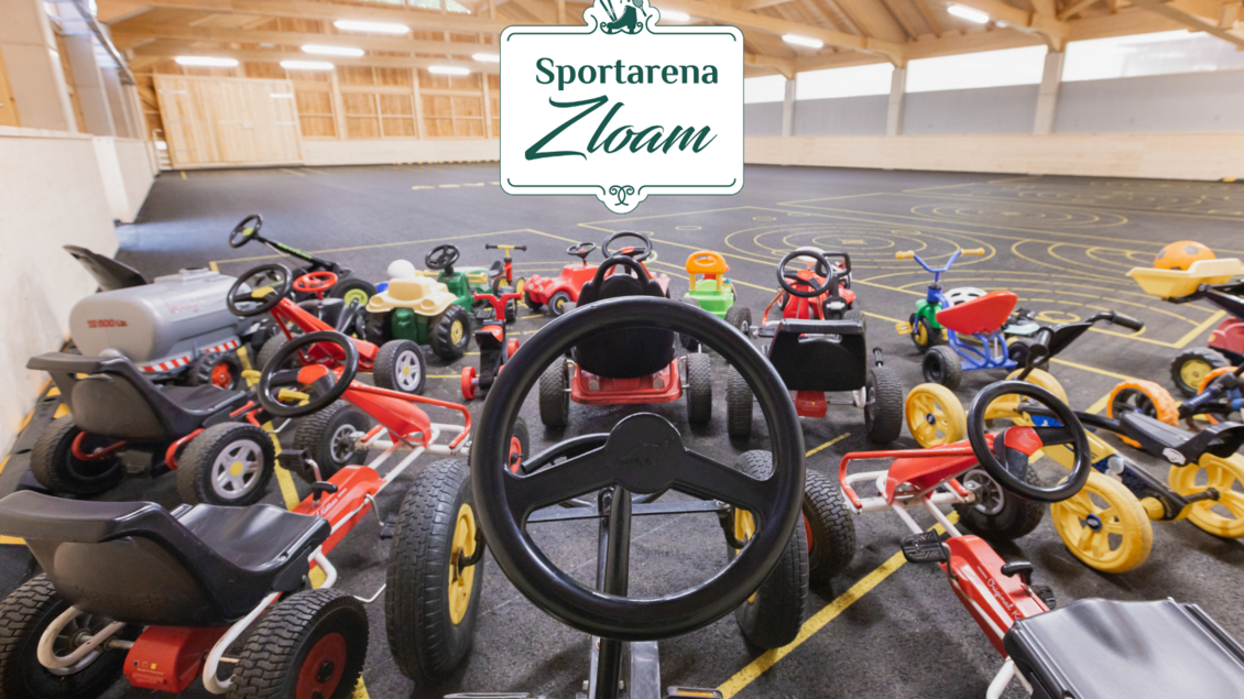 Eine Vielzahl von Kinderfahrzeugen steht in einer Sportarena. Die bunten Fahrzeuge sind auf einem schwarzen Boden angeordnet und laden zum Spielen ein.