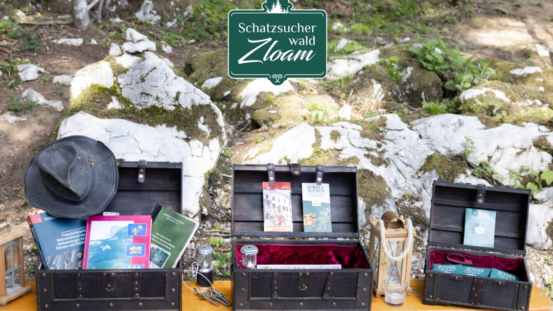 Drei Holzkisten stehen auf einem Tisch im Wald, gefüllt mit verschiedenen Broschüren und einem Hut. Im Hintergrund sind Steine und grüne Pflanzen zu sehen. | © Narzissendorf Zloam, www.zloam.at