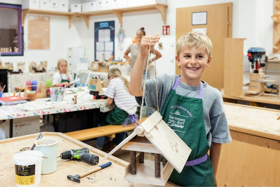 Ein junger Junge hält stolz ein selbstgebautes Vogelhaus in der Werkstatt. Im Hintergrund arbeiten andere Kinder an ihren Projekten. | © www.zloam.at