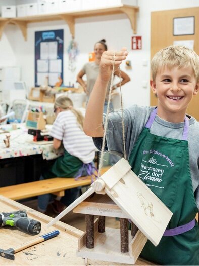 A young boy proudly holds a self-built birdhouse in the workshop. In the background, other children are working on their projects. | © www.zloam.at