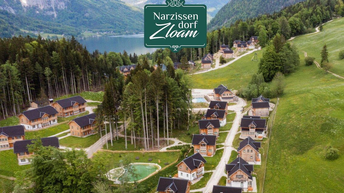 Ein malerisches Dorf mit Holzchalets inmitten von grünen Hügeln und Wäldern. Im Hintergrund ist ein klarer See und majestätische Berge zu sehen. | © Narzissendorf Zloam, www.zloam.at