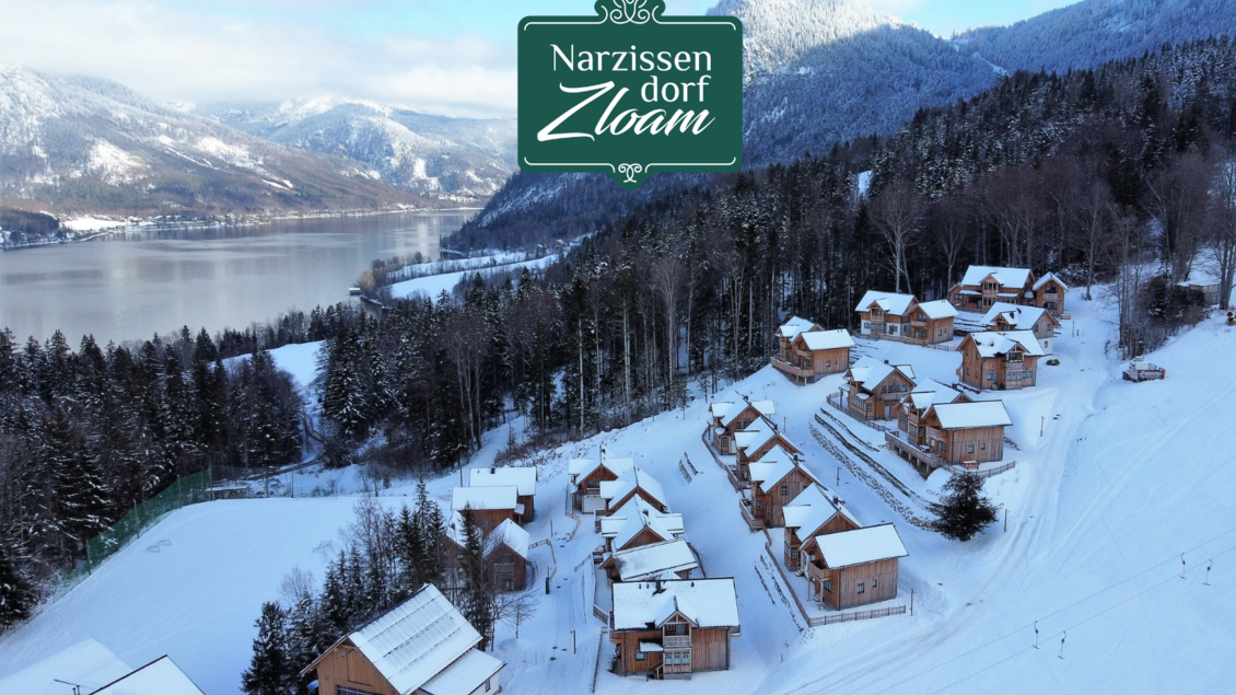 Ein winterliches Dorf mit gemütlichen Hütten im Schnee, umgeben von schneebedeckten Bäumen und Blick auf einen See. Die Landschaft ist malerisch und einladend. | © Narzissendorf Zloam, www.zloam.at