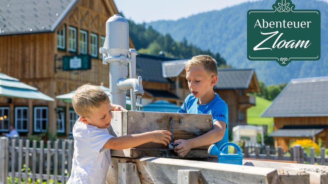 Zwei Kinder spielen miteinander an einem Spielplatz im Freien. Im Hintergrund sind Holzgebäude und eine malerische Landschaft zu sehen. | © www.zloam.at