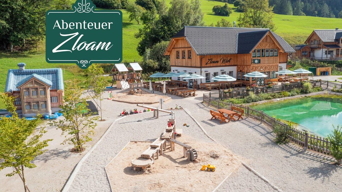 Ein schöner Spielplatz mit Sand und Spielgeräten. Im Hintergrund sind gemütliche Holzhäuser und ein Teich zu sehen. | © www.zloam.at