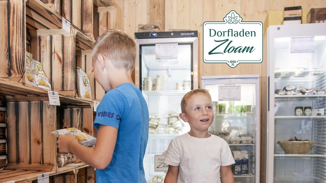 Ein Dorfshop mit Holzkisten voller Lebensmittel. Zwei Jungen, einer schaut neugierig zu und der andere wählt einen Snack aus. | © www.zloam.at