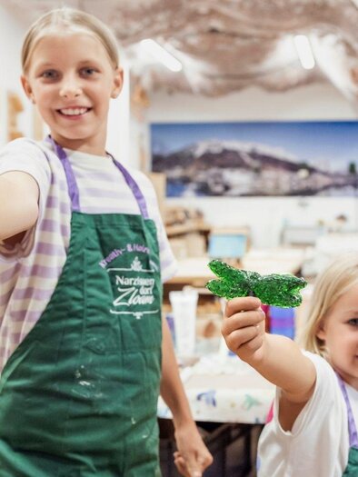 Two girls proudly hold their crafted creations in their hands. In the background, other children and adults can be seen in a creative studio. | © www.zloam.at