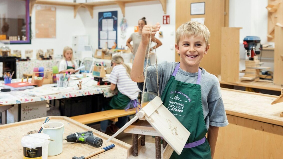 Ein Junge hält stolz ein selbstgebautes Vogelhaus in der Hand. Im Hintergrund arbeiten weitere Kinder an ihren Projekten in einem kreativen Raum. | © www.zloam.at