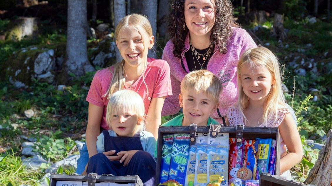 Eine Gruppe von fröhlichen Kindern steht vor einem kleinen Schatzkistchen im Freien. Sie lächeln in die Kamera und genießen den sonnigen Tag. | © www.zloam.at