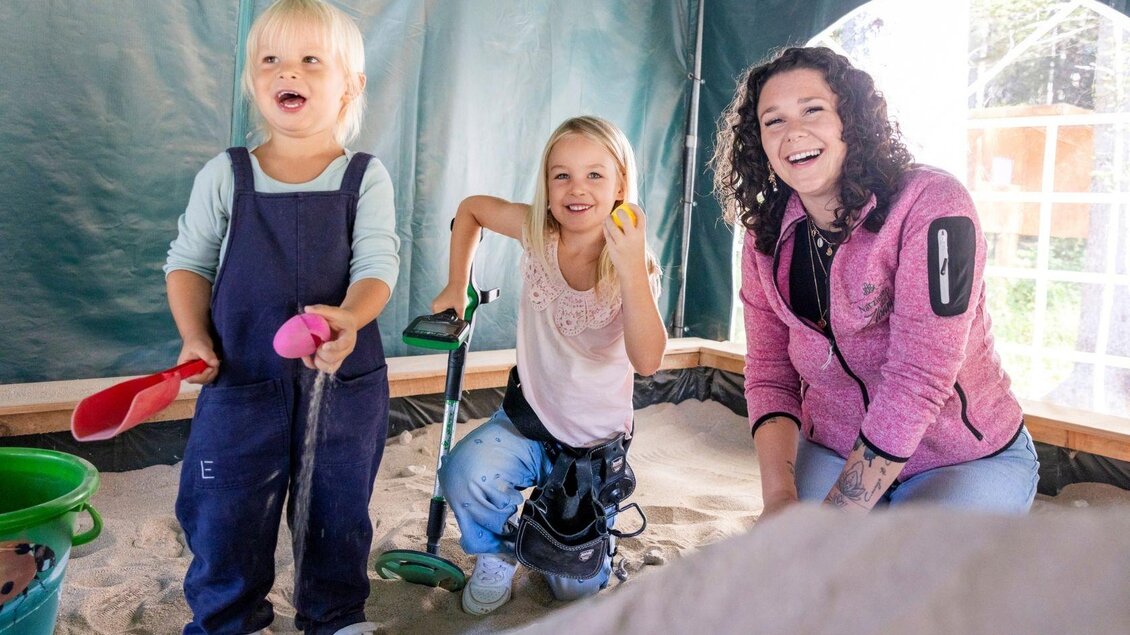 Zwei Mädchen spielen im Sandkasten und lachen fröhlich. Eine Frau steht neben ihnen und lächelt. | © www.zloam.at