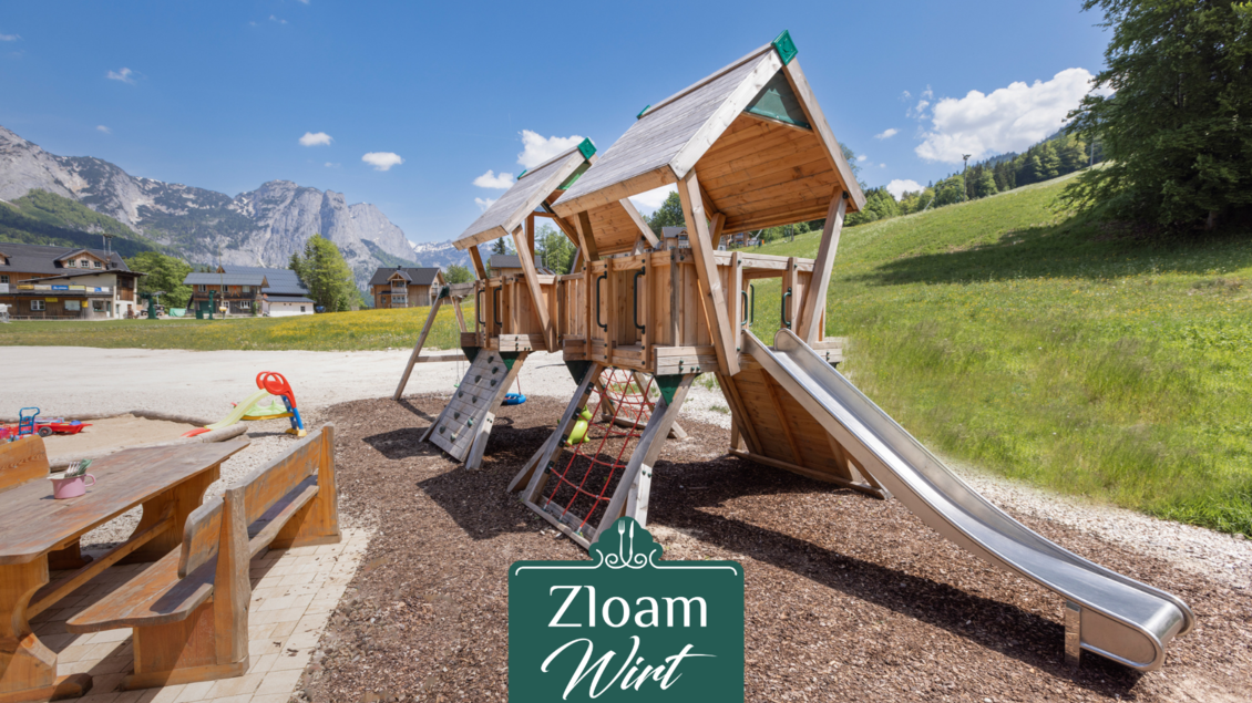 Ein schöner Spielplatz aus Holz mit einer Rutsche und Kletterelementen. Im Hintergrund sind grüne Wiesen und Berge zu sehen. | © Narzissendorf Zloam, www.zloam.at