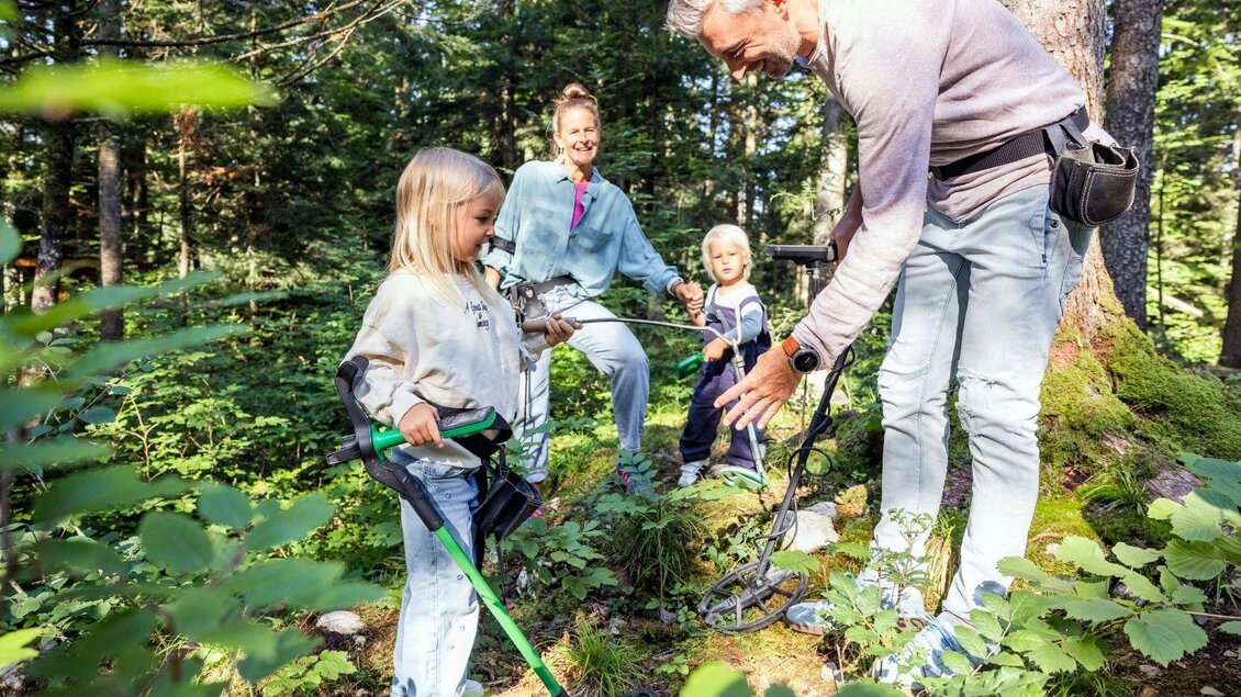 Eine Familie erkundet den Wald und benutzt einen Metalldetektor. Kinder und Erwachsene sind neugierig und beschäftigen sich mit der Natur.