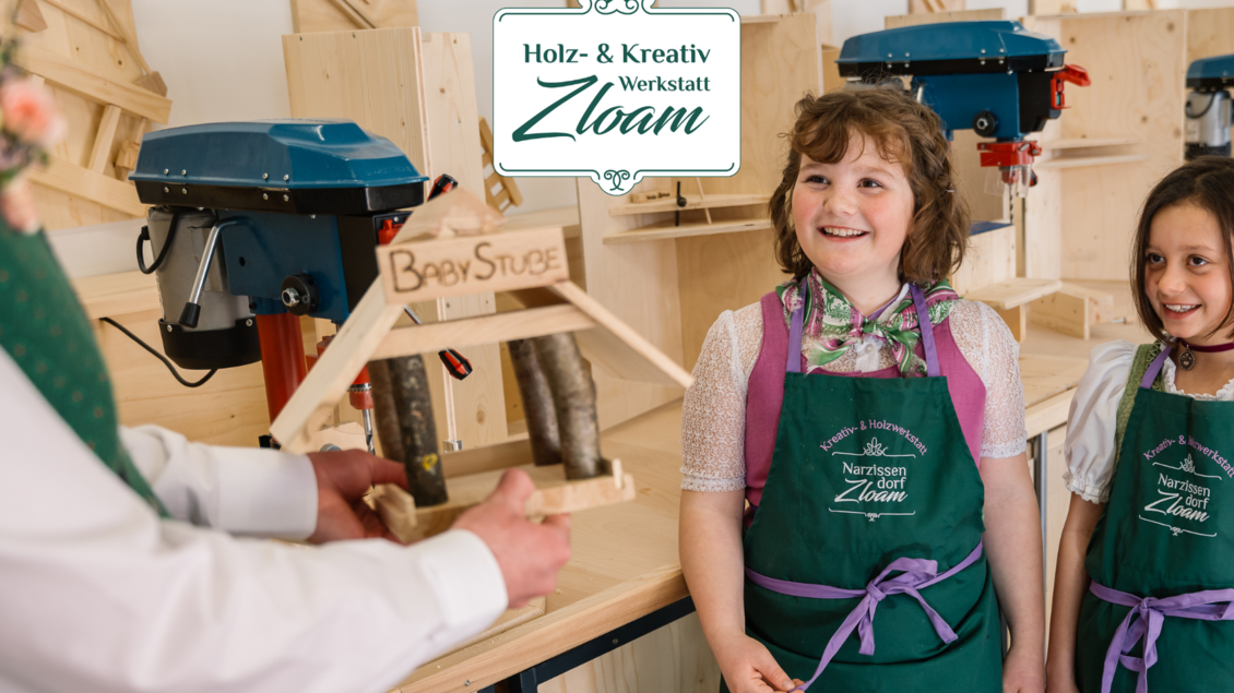 Zwei lächelnde Kinder in grünen Schürzen halten ein kleines Holzmodell in einer Werkstatt. Im Hintergrund sind Maschinen und eine freundliche Arbeitsatmosphäre zu sehen. | © Narzissendorf Zloam, www.zloam.at