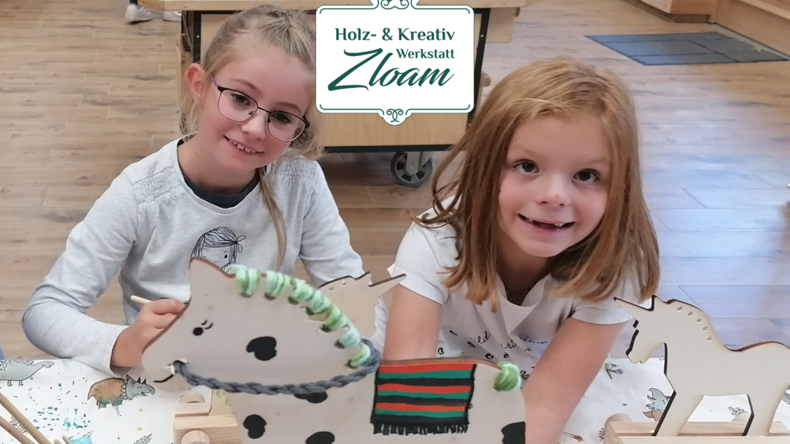Zwei junge Mädchen lächeln zufrieden und arbeiten kreativ an Holzfiguren in einer Werkstatt. Im Hintergrund sind Holzmaterialien und ein heller Raum zu sehen. | © Narzissendorf Zloam, www.zloam.at