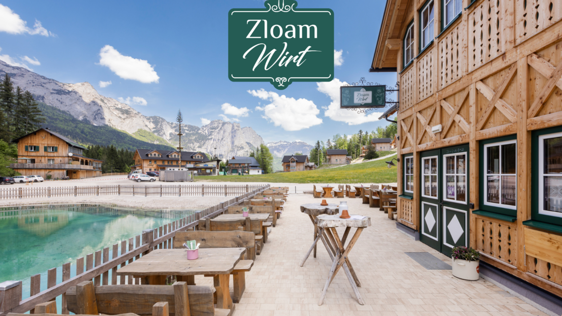 Eine einladende Terrasse mit Holzmöbeln am Wasser in einer malerischen Berglandschaft. Im Hintergrund sind charmante Hütten und majestätische Berge zu sehen. | © Narzissendorf Zloam, www.zloam.at