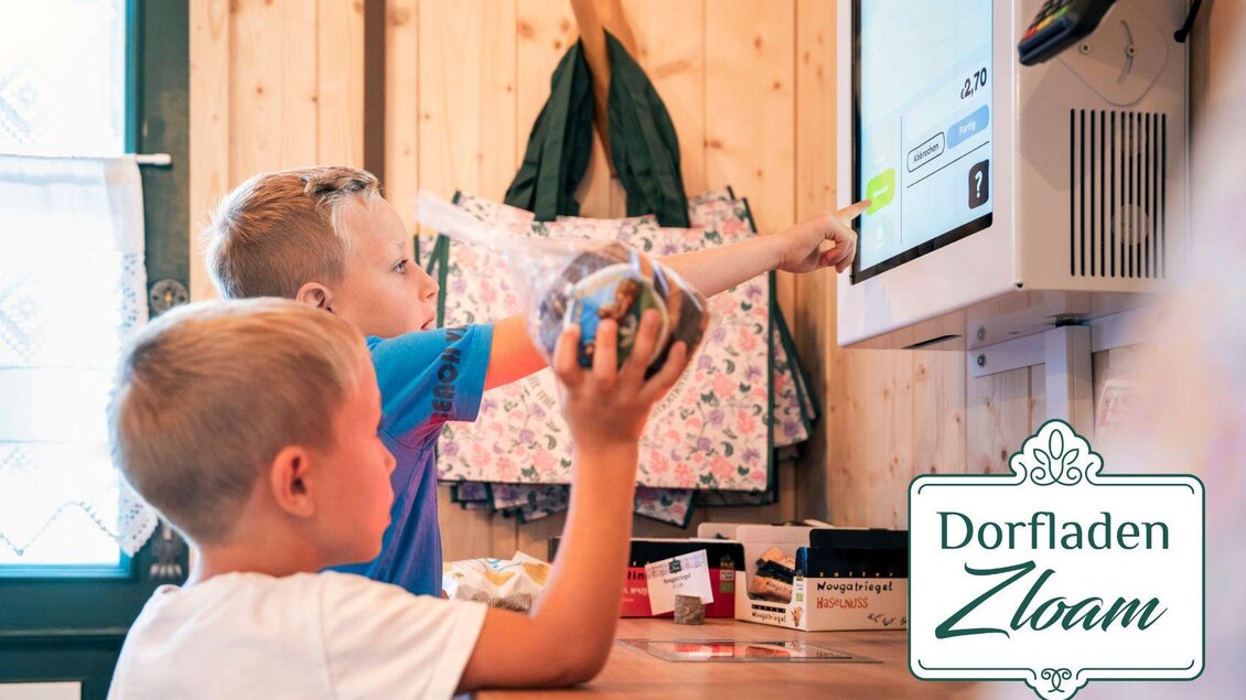 Zwei Kinder stehen an einem Tisch in einem Dorfladen und schauen auf einen Bildschirm. Eines der Kinder hält ein Glas in der Hand und zeigt auf den Bildschirm. | © www.zloam.at